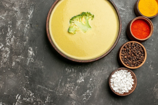 Horizontal view of creamy broccoli soup in a brown bowl and different spices on gray table