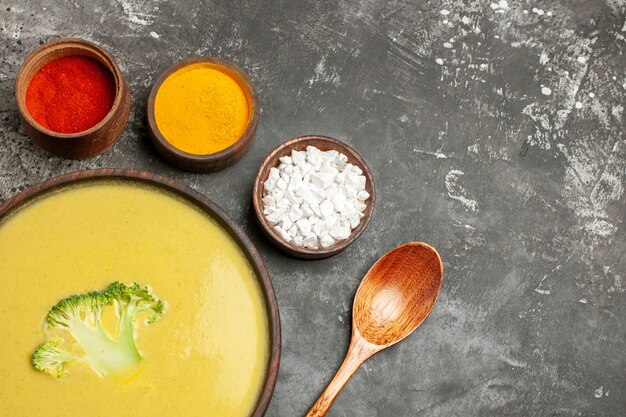 Horizontal view of creamy broccoli soup in a brown bowl and different spices on gray table