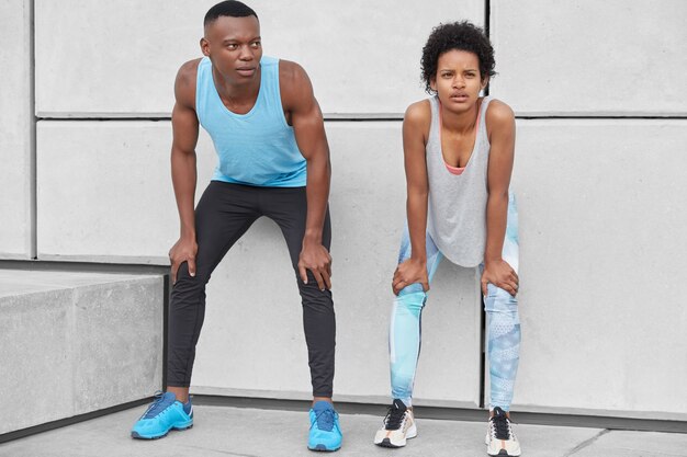 Horizontal view of fatigue woman and man lean at knees, stand closely to white wall, catch breath after intensive run, wear sneakers, leggings and t shirt, have contemplative expressions