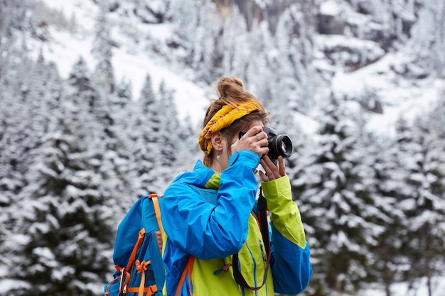 Free photo horizontal view of female hiker poses in snowy mountains, reaches top, makes photo with camera from hill