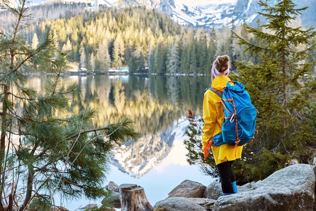 Horizontal view of female tourist enjoys tranquil remote mountain lake view, stands back to camera