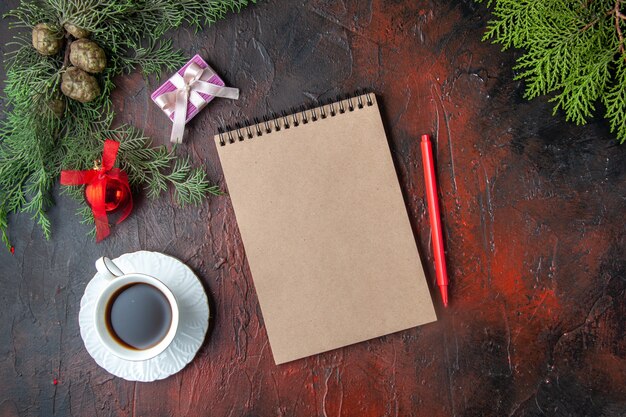 Horizontal view of fir branches a cup of black tea decoration accessories and gift next to notebook with pen on dark background