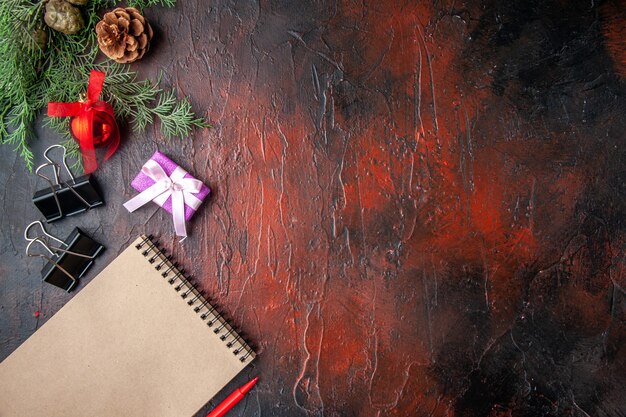 Horizontal view of fir branches decoration accessories and gift next to notebook with pen on dark background
