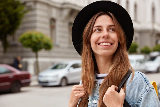 Horizontal view of glad smiling female pedestrian strolls across streets, looks away happily