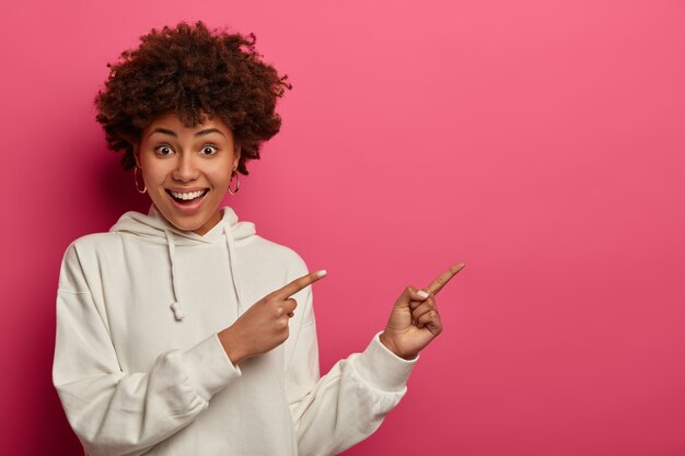 Horizontal view of glad young woman with cheerful expression shows direction aside, demonstrates awesome promo, shows way to cafe, suggests read banner, dressed in white hoodie. Advertisement