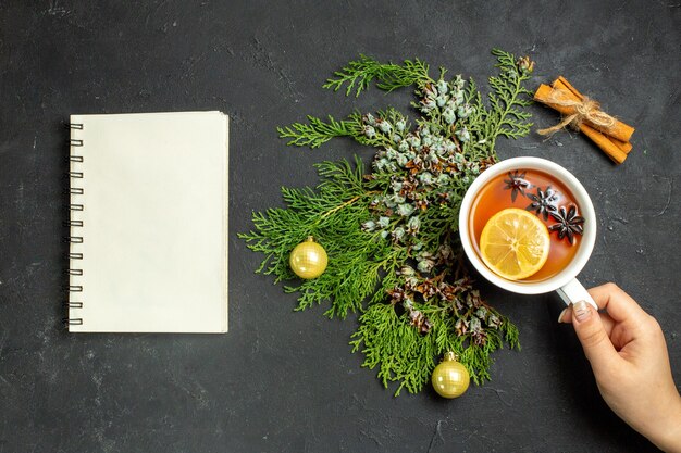 Horizontal view of hand holding a cup of black tea xsmas accessories and cinnamon limes and notebook on black background