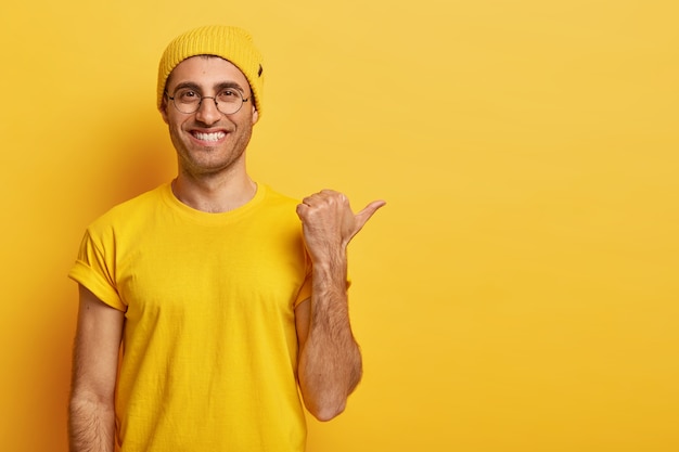 Horizontal view of happy delighted guy with toothy smile, points thumb away, has friendly look