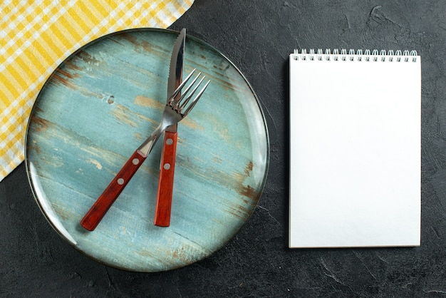 Horizontal view of meal cutlery in cross on a blue plate and yellow stripped towel next to notebook on dark surface