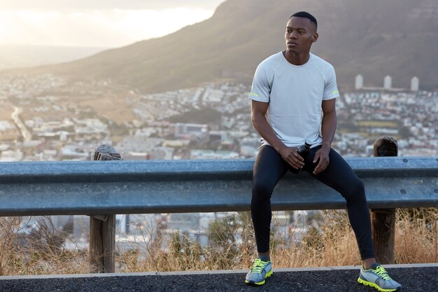 Horizontal view of motivated black male feels relaxed and pleased after jogging exercises, looks pensively aside, holds bottle with drink, stand against rock view with copy space on left side for text