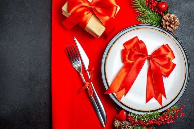 Горизонтальный вид новогоднего фона с красной лентой на обеденной тарелке, набор столовых приборов, украшения, аксессуары, еловые ветки, рядом с подарком на красной салфетке