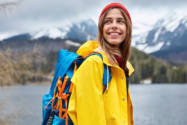 Horizontal view of optimistic European female tourist looks happily aside, enjoys walking near mountain lake, admires beautiful scenery. People