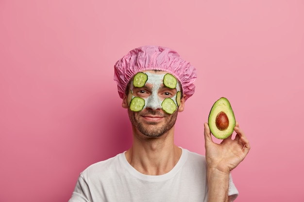 Horizontal view of pleased young Caucasian man shows half of avocado, enjoys nourishing clay mask on face, has stubble, dressed in white t shirt