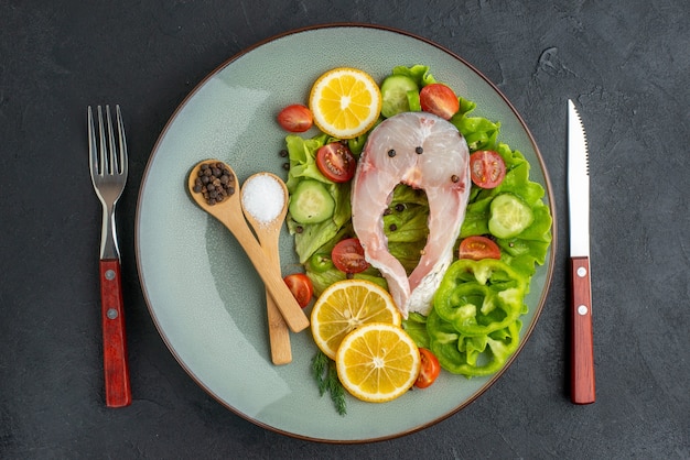 Horizontal view of raw fish and fresh chopped vegetables lemon slices spices on a gray plate and cutlery set on black distressed surface