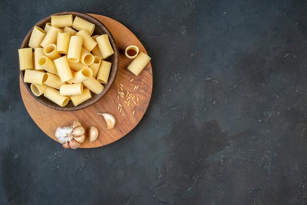 Horizontal view of raw pastas in a brown bowl garlics rice on wooden board on the right side on black background