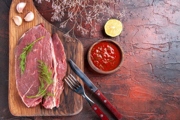 Horizontal view of red meat on wooden cutting board and ketchup in small bowl fork and knife lemon on dark background