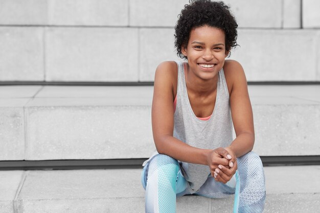 Horizontal view of relaxed pleased dark skinned woman with curly haircut, has toothy smile, shows white teeth, being in good shape as goes in for sport every day, rests at city stairs with free space