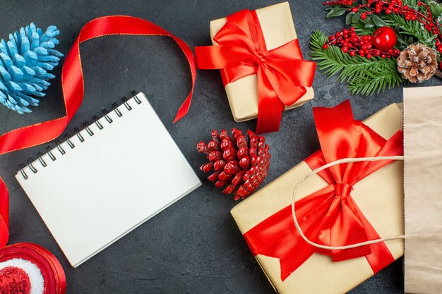 Horizontal view of a roll of red ribbon conifer cones and gift fir branches next to notebook on dark background