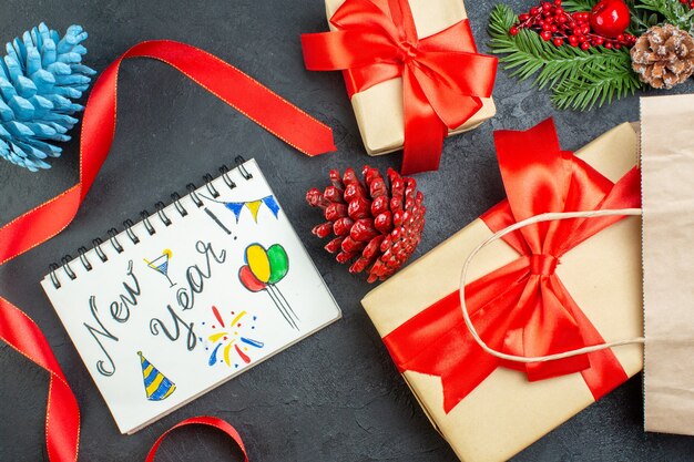 Horizontal view of a roll of red ribbon conifer cones and gift fir branches notebook with new year drawings on dark table