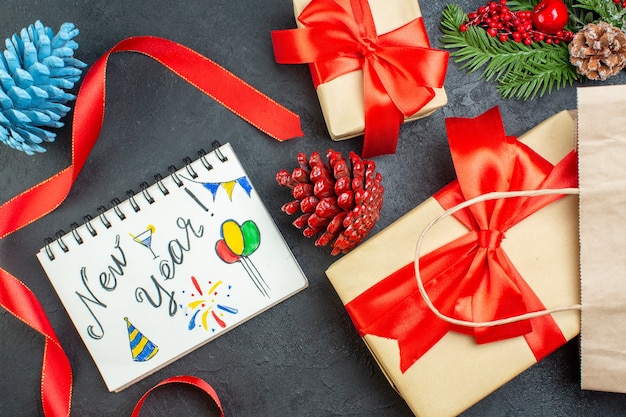 Horizontal view of a roll of red ribbon conifer cones and gift fir branches notebook with new year drawings on dark table