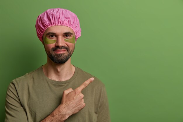 Horizontal view of satisfied unshaven man wears pink bath hat, casual t shirt, has eye skin treatment, applies collagen pads for reducing fine lines, points at blank space, advertises some product