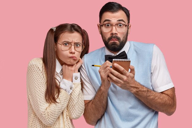 Horizontal view of surprised male geek and his beautiful girlfriend with pony tails, wears thick lenses spectacles, make records in small notepad