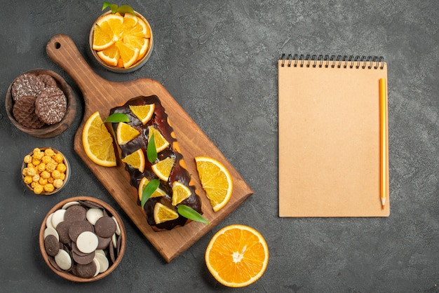 Horizontal view of tasty cakes cut oranges with biscuits and notebook on cutting board on dark table