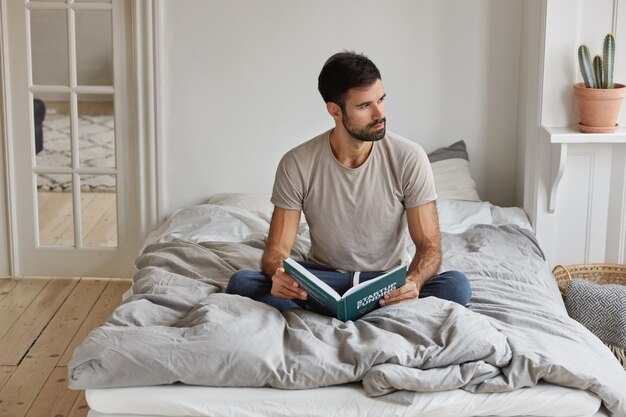 Horizontal view of thoughtful bearded man holds book, sits crossed legs on comfortable bed