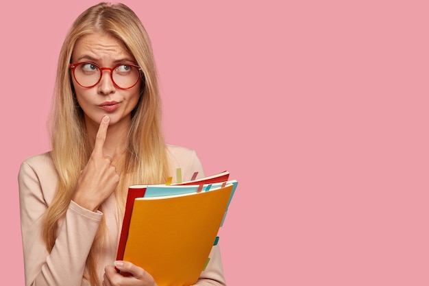 Horizontal view of thoughtful schoolgirl purses lips, keeps index finger on chin, wears round glasses