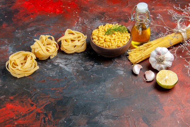 Horizontal view of uncooked three portions of spaghetti and butterfly pastas in a brown bowl and green onion lemon garlic oil bottle on mixed color table