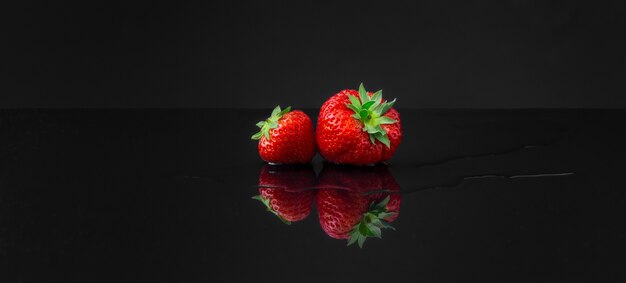 Horizontal wide-angle shot of two red strawberries on a black reflecting surface