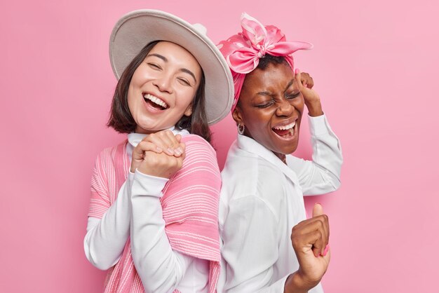 Horizotal shot of cheerful young female models have fun together dance with rhythm of music laugh happily stand back to each other isolated over pink background foolish around. Happiness concept.