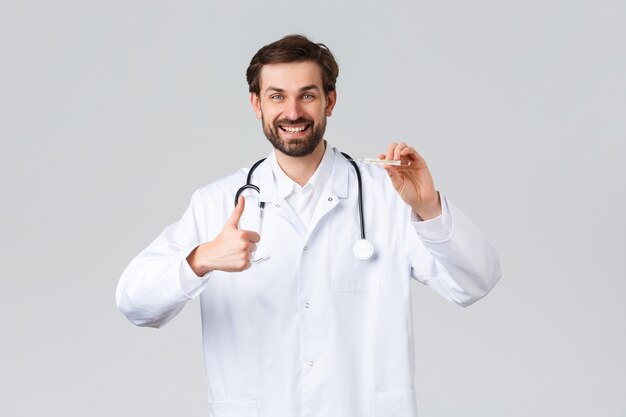 Hospital, healthcare workers, covid-19 treatment concept. Handsome friendly doctor in white scrubs, with stethoscope showing thumbs-up as measure patient temperature perfect fine, show thermometer.