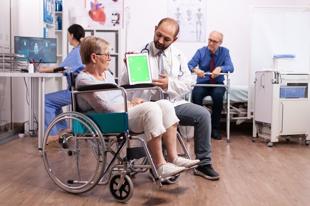 Hospital staff consulting handicapped senior woman sitting in wheelchair holding tablet pc with green screen