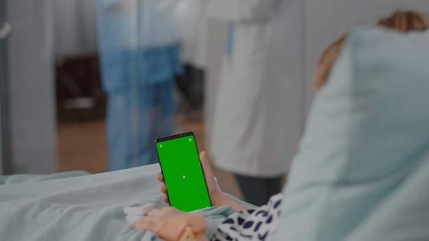 隔離されたディスプレイとモックアップグリーンスクリーンクロマキースマートフォンを見てベッドで休んでいる入院中の小さな子供。病棟での回復相談中に手術を受けた病気の子供