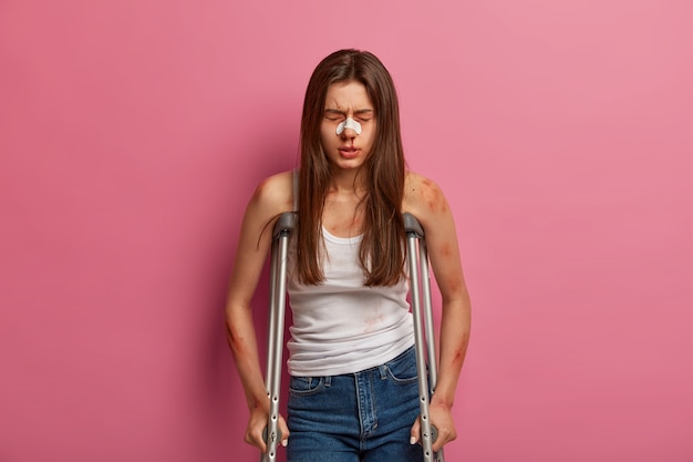 Hospitilized woman has rehabilitation period after serious accident, various fractures, poses on crutches, suffers from serious spine disease, injured after car crash, has broken bleeding nose