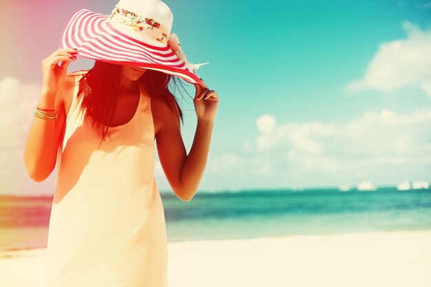 Hot beautiful woman in colorful sunhat and dress walking near beach ocean on hot summer day on white sand