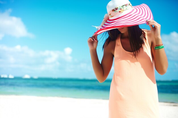 Hot beautiful woman in colorful sunhat and dress walking near beach ocean on hot summer day on white sand