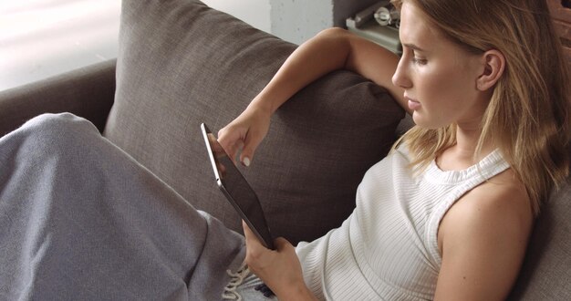 Hot blond model reading on her tablet while sitting on a dark gray couch covered with a soft throw