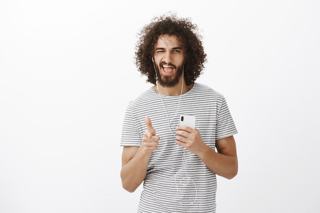 Hot confident curly-haired boyfriend in striped t-shirt, holding smartphone, pointing with finger gun at front