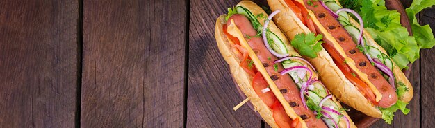 Hot dog with  sausage, cucumber, tomato and lettuce on dark wooden table. Summer hotdog. Top view