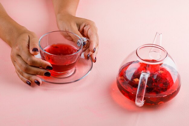 Hot drink concept with teapot on pink table woman holding glass cup of tea.