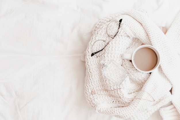 Hot drink, necklace, eyeglasses and perfume on sweater placed on bedsheet