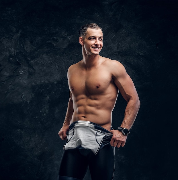 Hot sexy fit man is taking off his wetsuit in dark photo studio.