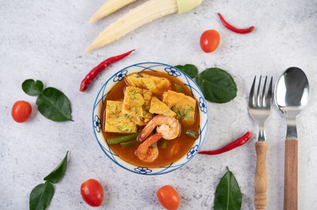 Hot and sour soup with cha-om, egg, and shrimp in a white bowl, with chili and kaffir lime leaves on white surface.