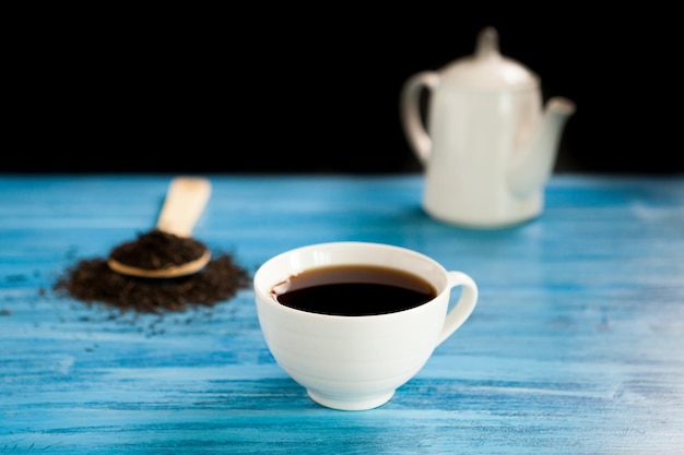 Foto gratuita tè caldo accanto a un cucchiaio con foglie di tè sul bordo blu vintage su sfondo nero