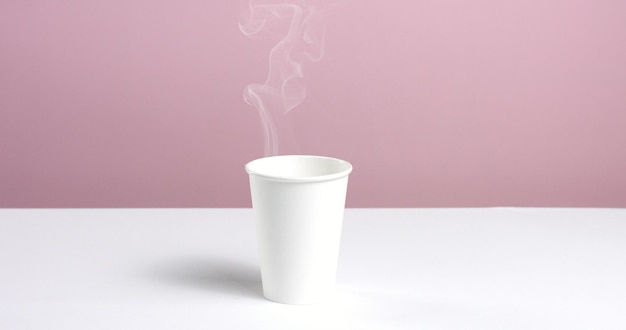 Foto gratuita acqua calda o caffè versato in una semplice tazza di carta senza etichetta sul tavolo bianco contro la parete rosa in aumento del vapore