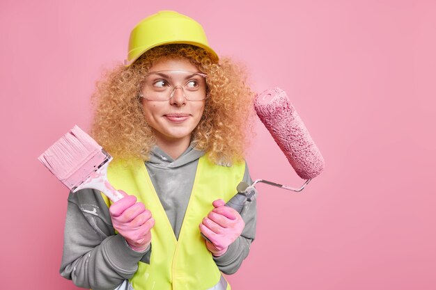 House construction and repair service. Thoughtful professional woman constructor with curly bushy hair wears hardhat and transparent glasses safety helmet gloves uniform poses against pink wall