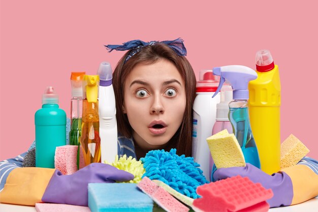 Household concept. Stupefied dark haired woman stares through bottles of dishwashing liquids, uses detergent for microvave oven and stoves