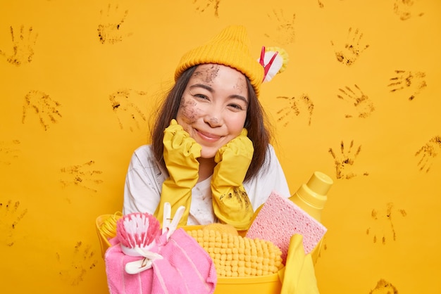 家事​の​概念​満足している​アジア​の​女性​は​疲れ​を​感じます​が​、​あご​の​下​で​満足している​手​は​帽子​を​かぶっていて​、​ゴム​手袋​は​黄色​の​背景​の​上に​隔離された​洗濯物​と​洗剤​で​バスケット​に​寄りかかっています