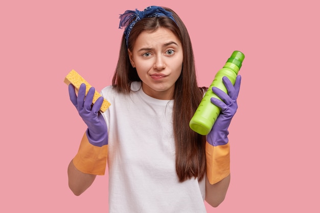 Концепция уборки и уборки. Несчастная смущенная молодая женщина-уборщик с длинными волосами, держит губку и зеленую бутылку с моющим средством, носит резиновые перчатки для защиты рук, выполняет домашние дела